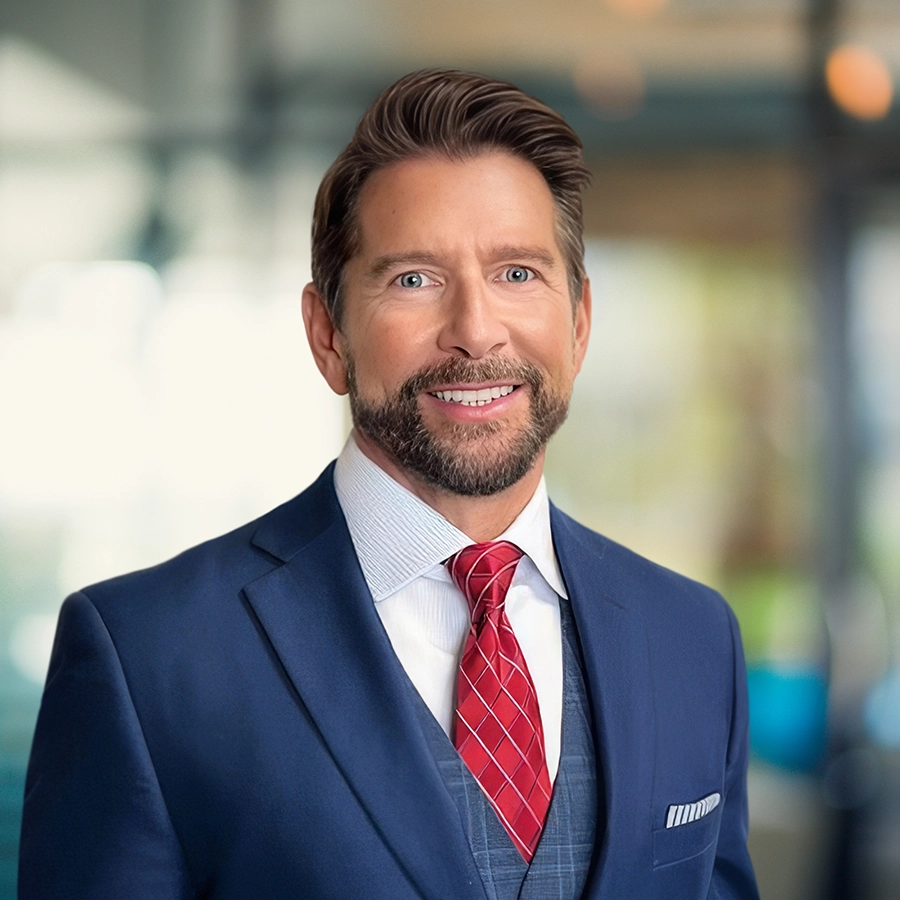 A man in a blue suit, white shirt, and red tie stands indoors, smiling at the camera with a blurred office background.