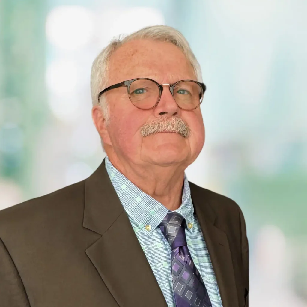 An older man with gray hair, glasses, and a mustache is wearing a suit, dress shirt, and patterned tie, standing against a blurred light background.
