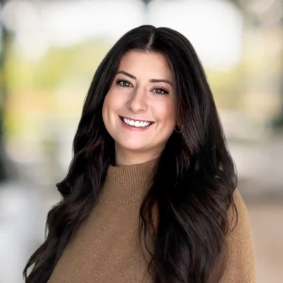 A woman with long dark hair wearing a brown sweater smiles at the camera with a blurred outdoor background.