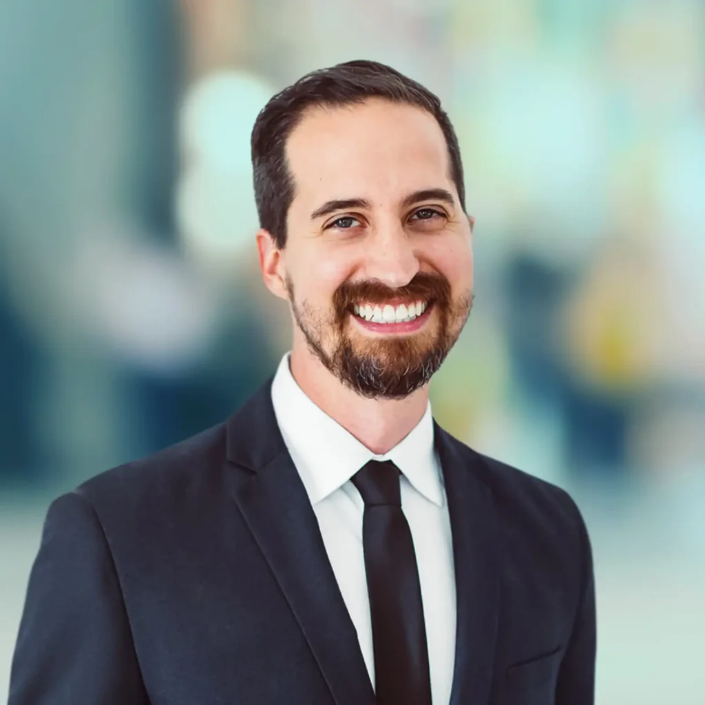 A man with dark hair and a beard, wearing a black suit, white shirt, and black tie, smiles at the camera against a blurred indoor background.