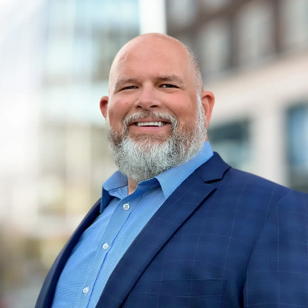 A man with a trimmed white beard wearing a blue suit jacket and a light blue shirt smiles at the camera outdoors, with blurred buildings in the background.