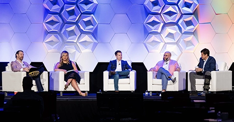 Five people sit on stage in armchairs during a panel discussion, with a geometric patterned backdrop behind them.
