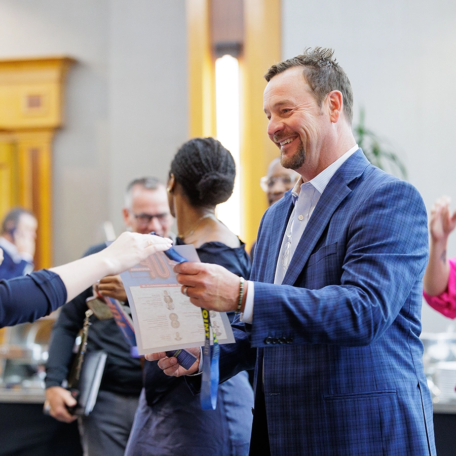 A man in a blue jacket smiles as he receives a booklet from someone at an indoor event, with other people in the background.