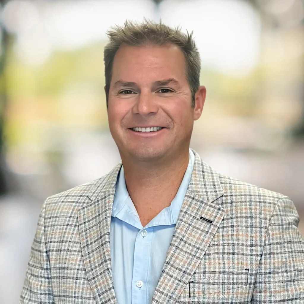 A man with short light brown hair, wearing a light blue shirt and a plaid blazer, smiles at the camera with a blurred background.