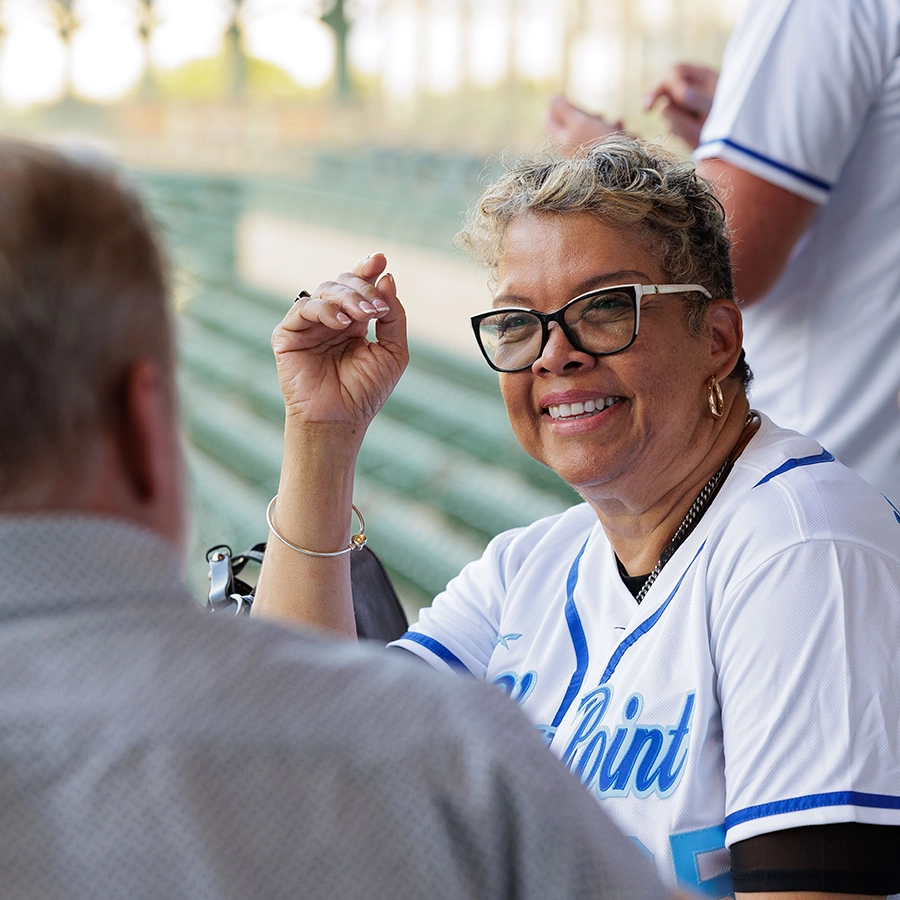 A woman in a white baseball jersey and glasses smiles while sitting and talking to a man in a stadium with empty green seats.