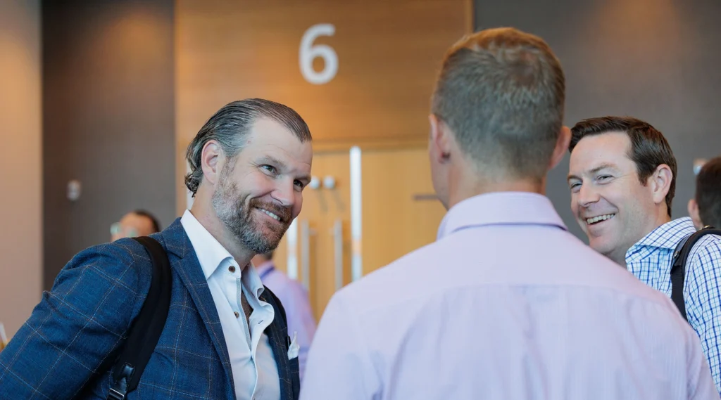 Three men in business attire have a conversation in front of a door labeled with the number 6.