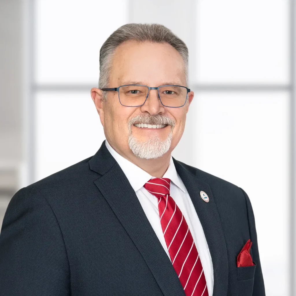 A middle-aged man with gray hair, glasses, and a goatee is wearing a dark suit, white shirt, red striped tie, and pocket square, standing in front of a bright window.