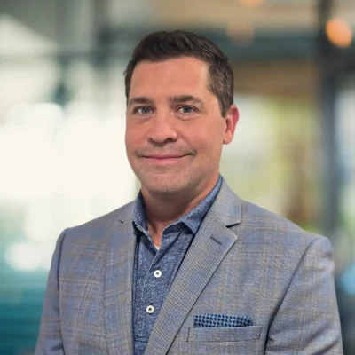 A man wearing a blue checked blazer and blue shirt stands in an indoor setting with a blurred background.