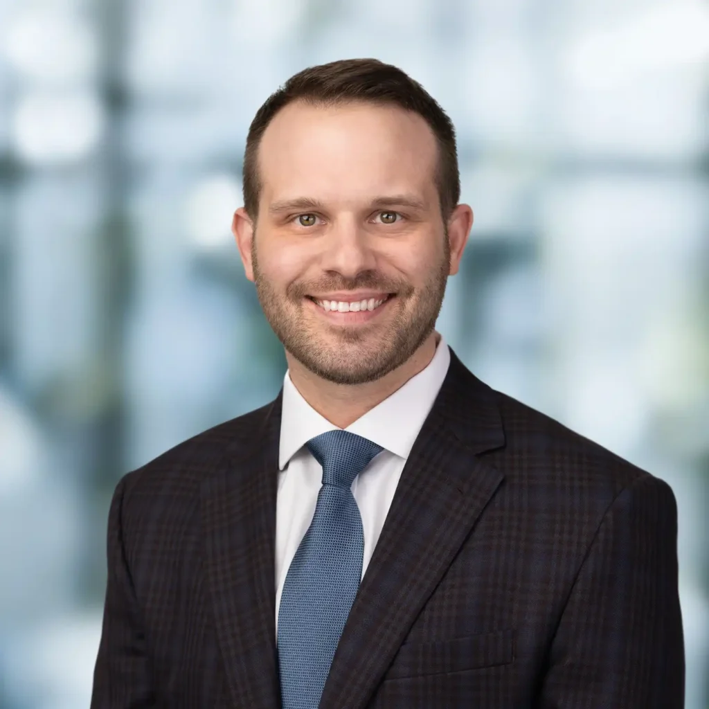 A man in a checked dark suit, light blue shirt, and blue tie smiles at the camera against a blurred office background.