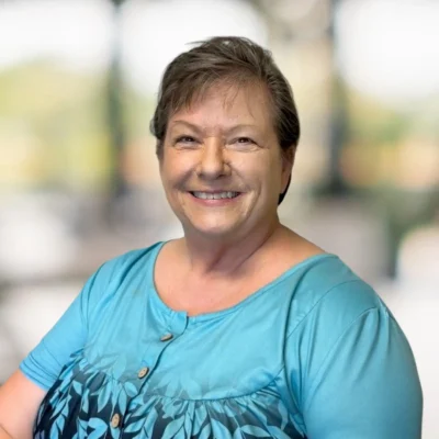 A woman with short brown hair smiles at the camera, wearing a turquoise top with a leaf pattern, against a blurred outdoor background.
