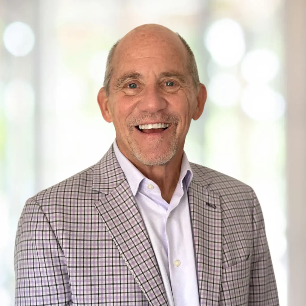 A smiling older man in a light plaid blazer and white shirt stands in front of a softly blurred background.