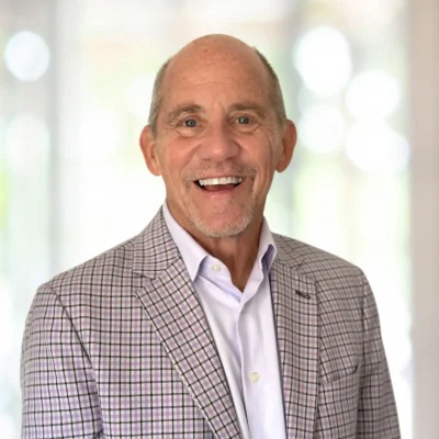 A smiling older man in a light plaid blazer and white shirt stands in front of a softly blurred background.