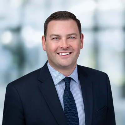 A man in a dark suit and tie smiles at the camera against a blurred office background.