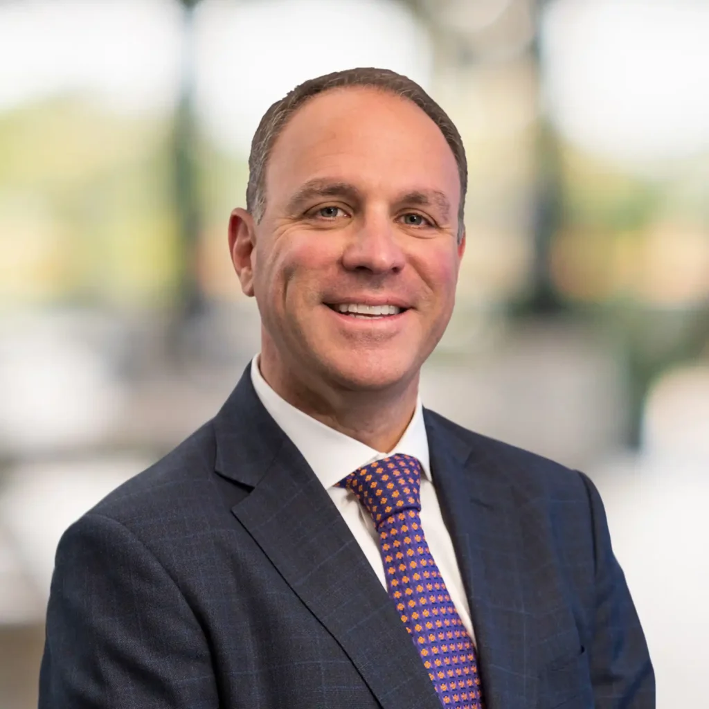 A man in a navy suit, white shirt, and patterned tie smiles at the camera with a blurred indoor background.