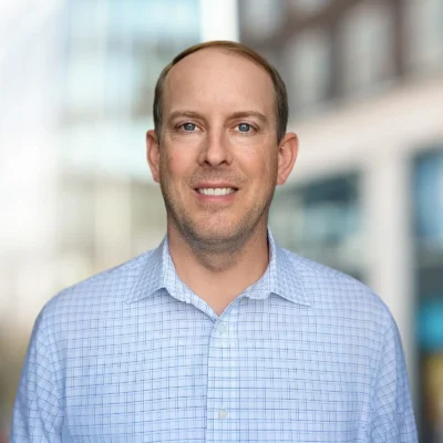 A man wearing a light blue checkered dress shirt stands outdoors with blurred buildings in the background.