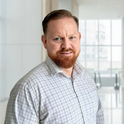 A man with red hair and a beard wearing a grey plaid shirt stands indoors in front of large windows with natural light.