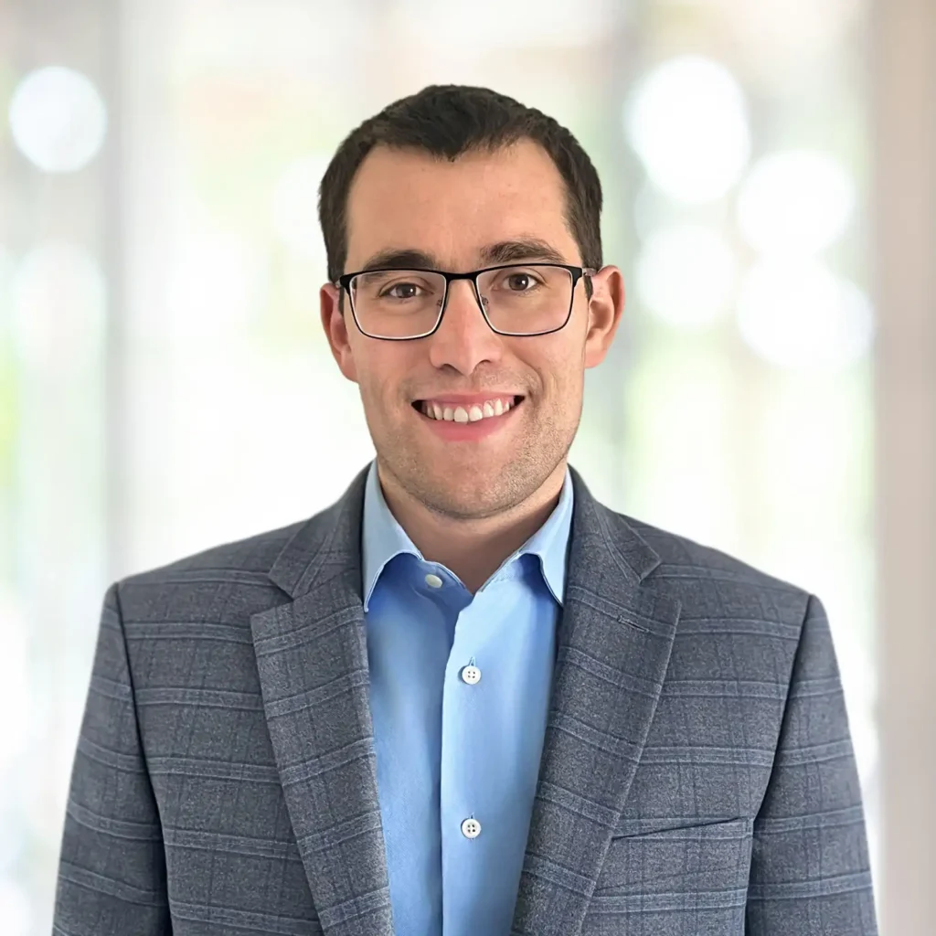 A man wearing glasses, a blue dress shirt, and a checked grey blazer smiles at the camera against a blurred, light background.
