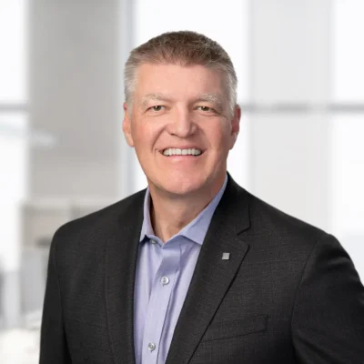 A middle-aged man in a dark suit jacket and light blue shirt smiles at the camera in a bright, modern office setting.