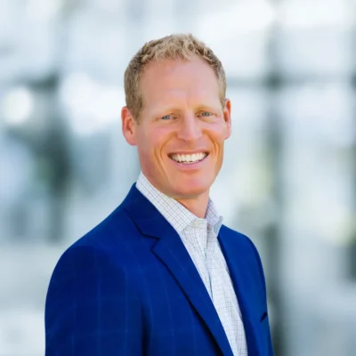 A man with short blond hair wearing a blue suit jacket and checkered shirt smiles while standing in front of a blurred background.