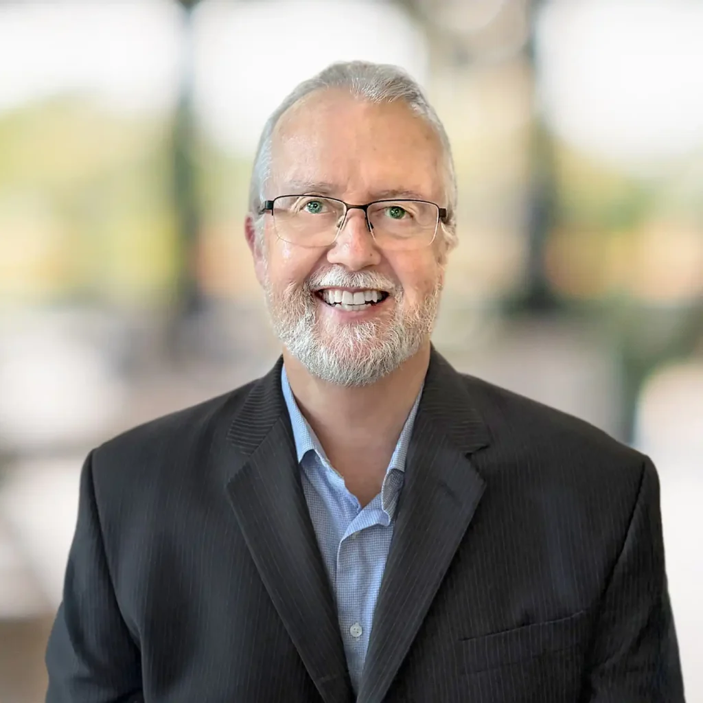 A man with gray hair, glasses, and a beard is smiling, wearing a dark suit jacket and blue shirt, with a blurred background.