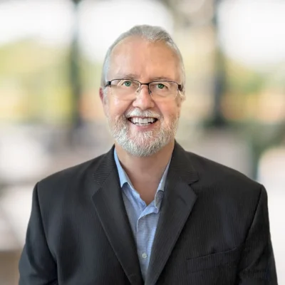 A man with gray hair, glasses, and a beard is smiling, wearing a dark suit jacket and blue shirt, with a blurred background.