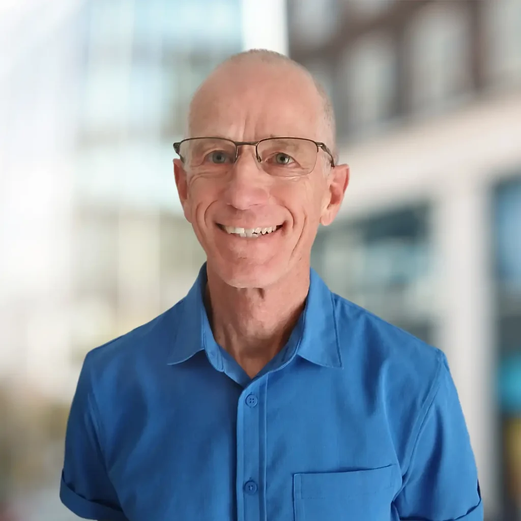 A smiling older man wearing glasses and a blue collared shirt stands in front of a blurred cityscape background.