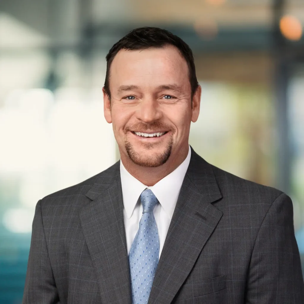 Man in a gray suit and light blue tie smiling at the camera, with a blurred office background.