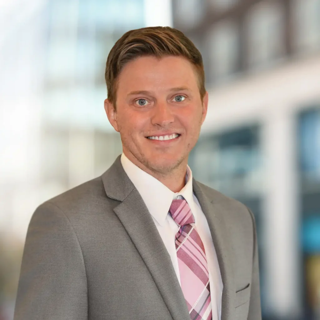 A man in a gray suit, white shirt, and pink striped tie is smiling at the camera with a blurred cityscape background.
