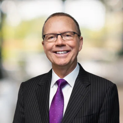 A man wearing glasses, a black pinstripe suit, white shirt, and purple tie is smiling at the camera with a blurred background.