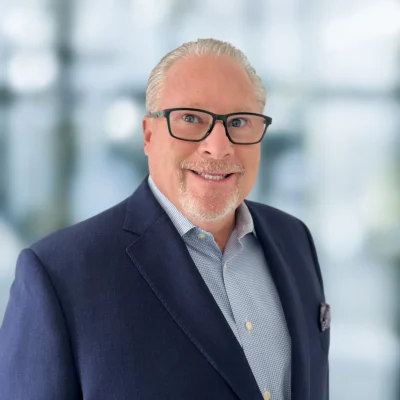 A middle-aged man with gray hair, glasses, and a neatly trimmed beard wearing a navy suit jacket and light blue shirt, standing in front of a blurred background.