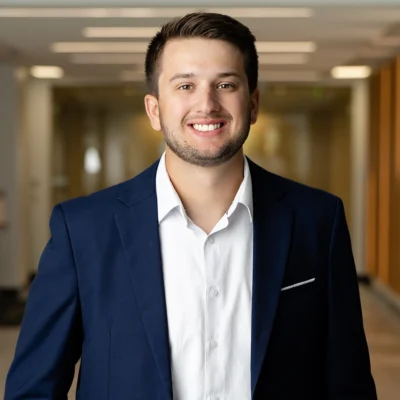 A young man in a navy suit and white shirt stands indoors, smiling at the camera in a well-lit hallway.