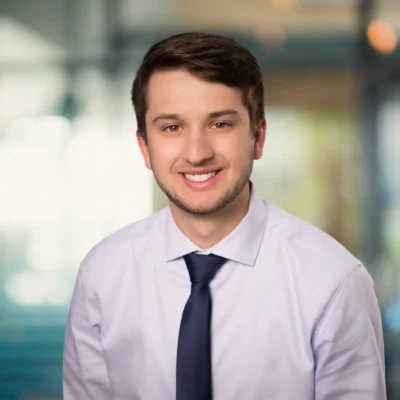 A young man with short brown hair, wearing a white shirt and dark tie, smiles at the camera in an office setting with a blurred background.