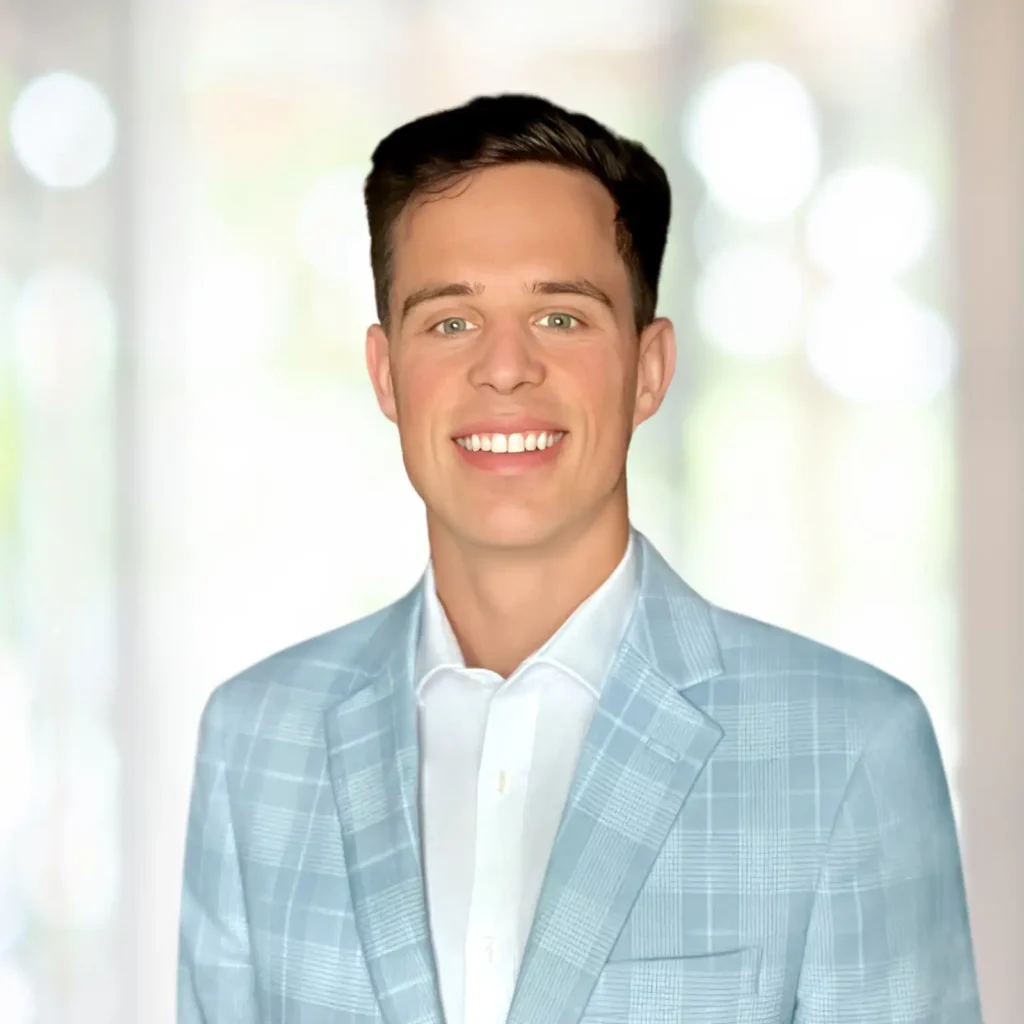 A man in a light blue plaid suit jacket and white shirt smiles at the camera against a blurred indoor background.
