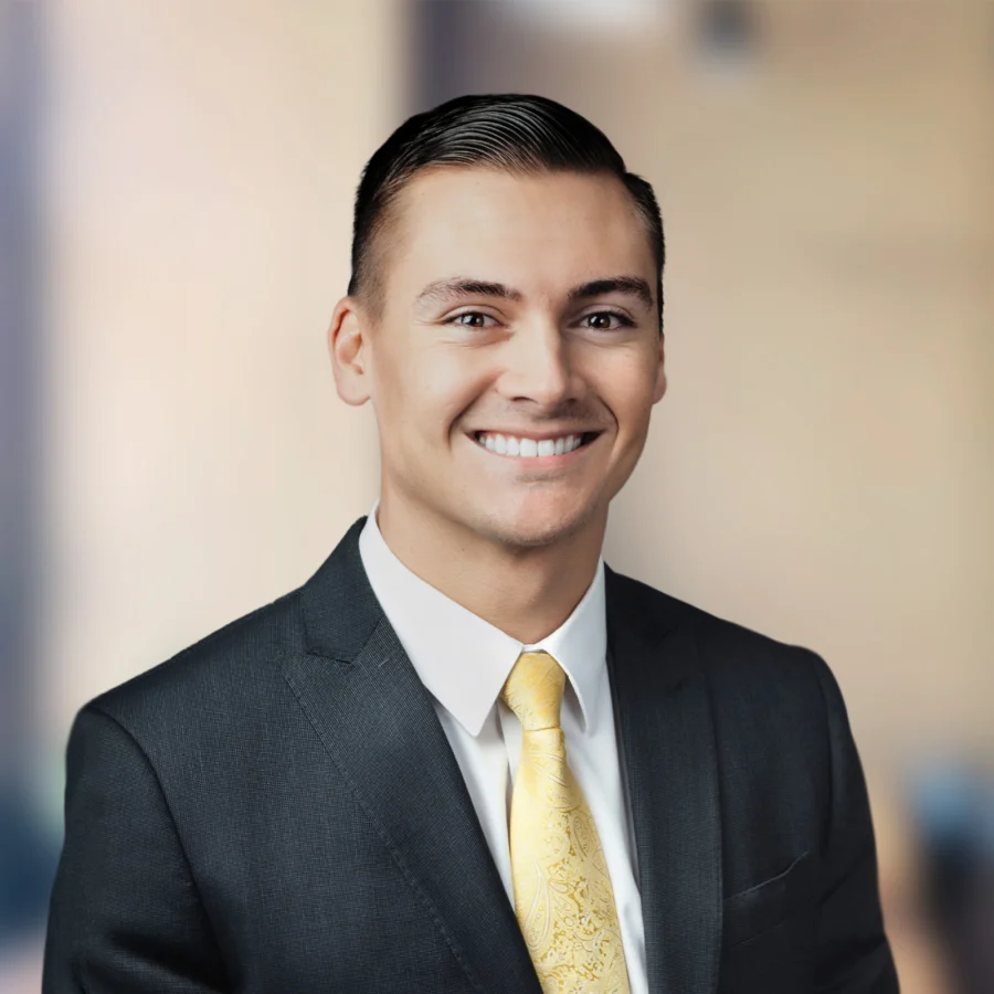 A man in a dark suit and yellow patterned tie smiles at the camera, standing in front of a blurred indoor background.