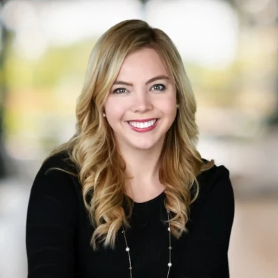A woman with long blonde hair, wearing a black top and necklace, smiles at the camera with a blurred outdoor background.
