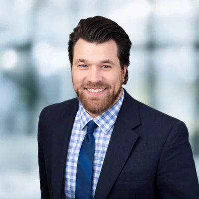 A man with dark hair and a beard, wearing a navy suit, blue checkered shirt, and blue tie, smiles at the camera against a blurred indoor background.