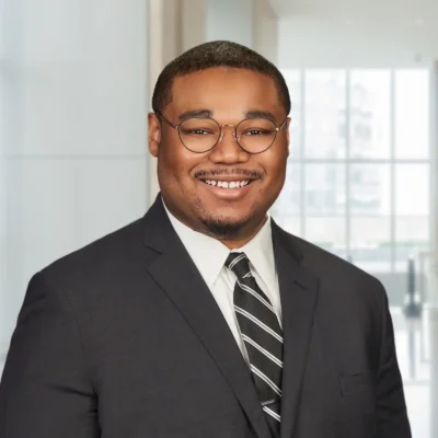 A man wearing glasses, a dark suit, white shirt, and striped tie smiles in a well-lit office setting with large windows in the background.