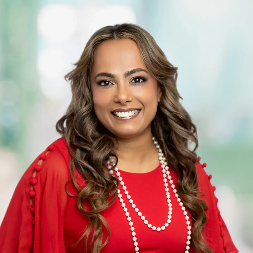 A woman with long wavy hair smiles at the camera. She is wearing a red outfit and layered pearl necklaces, with a blurred light background behind her.