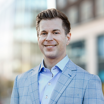 Man in a light blue checked suit jacket and light blue shirt standing outdoors with blurred modern buildings in the background.
