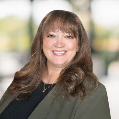 A woman with straight brown hair and bangs, wearing a dark blazer and necklace, smiles at the camera against a blurred background.