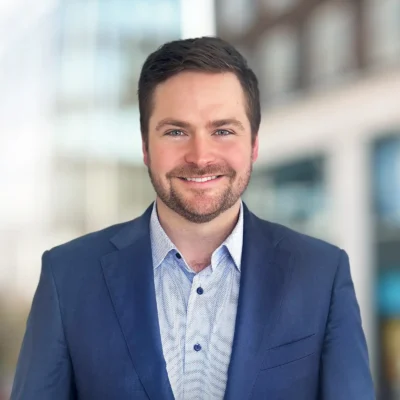 A man with short brown hair and a beard, wearing a blue suit jacket and light blue shirt, stands outdoors in front of a blurred city background.