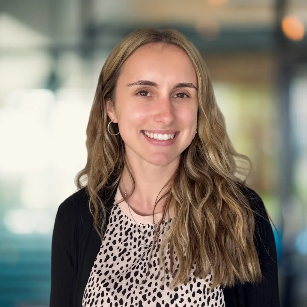A woman with long wavy blonde hair, wearing a black cardigan and a light-colored patterned top, smiles in an indoor setting with a blurred background.
