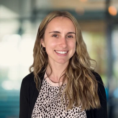 A woman with long wavy blonde hair, wearing a black cardigan and a light-colored patterned top, smiles in an indoor setting with a blurred background.