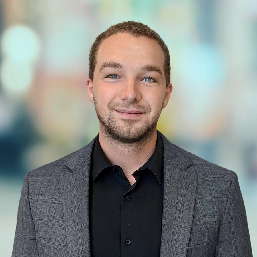 A man with short hair and a trimmed beard wears a gray plaid suit jacket over a black shirt, standing in front of a blurred background.