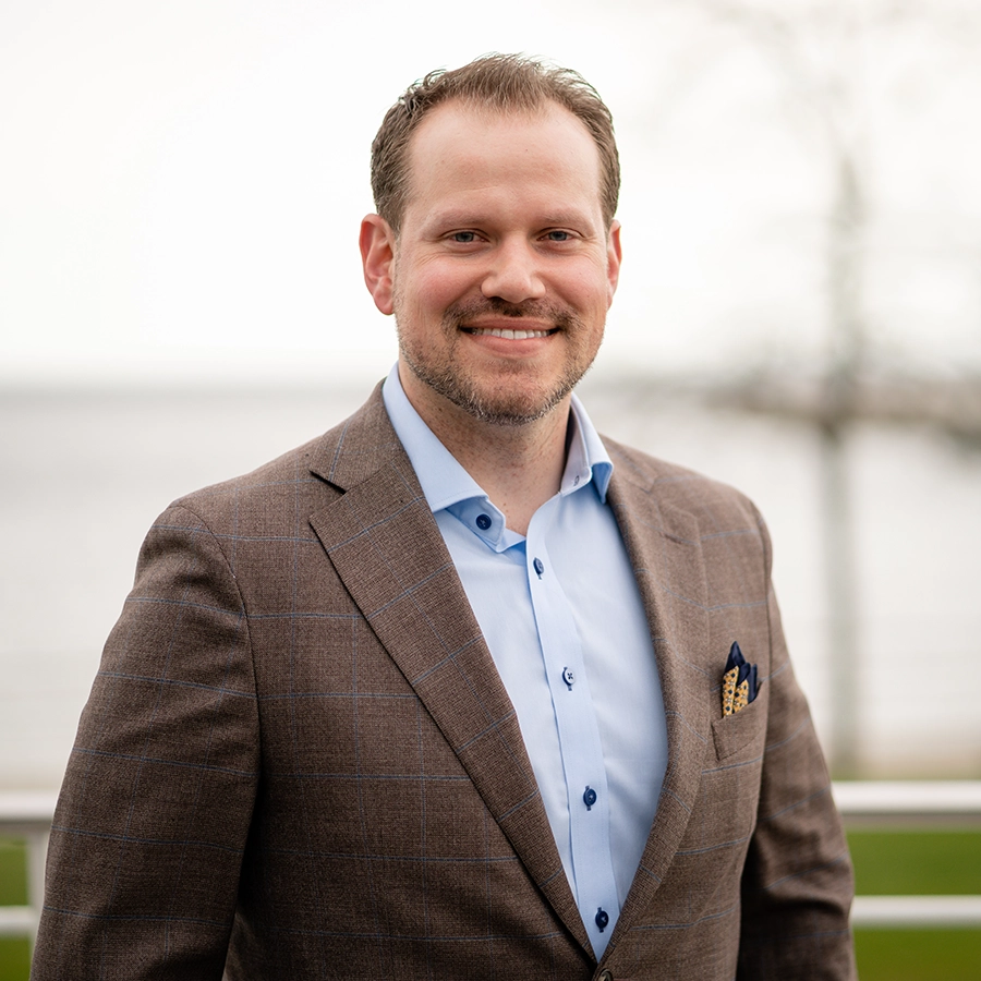 A man in a brown plaid suit jacket and light blue shirt stands smiling outdoors with a blurred background of water and trees.