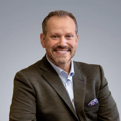 A man with short hair and a beard is smiling, wearing a dark blazer over a light shirt, sitting against a plain grey background.
