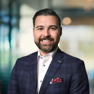 A man with short dark hair and a beard, wearing a checked suit jacket, white shirt, and red patterned pocket square, smiles in a modern indoor setting.