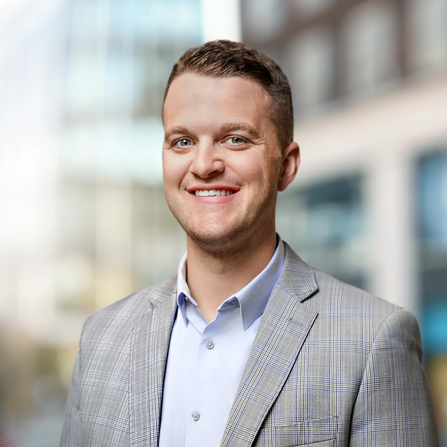 A man in a light gray plaid suit jacket and a light blue shirt is smiling at the camera with a blurred cityscape in the background.
