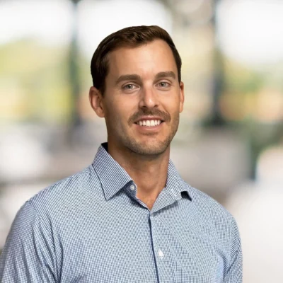 A man with short brown hair and a mustache, wearing a light blue checkered shirt, smiles at the camera against a blurred indoor background.