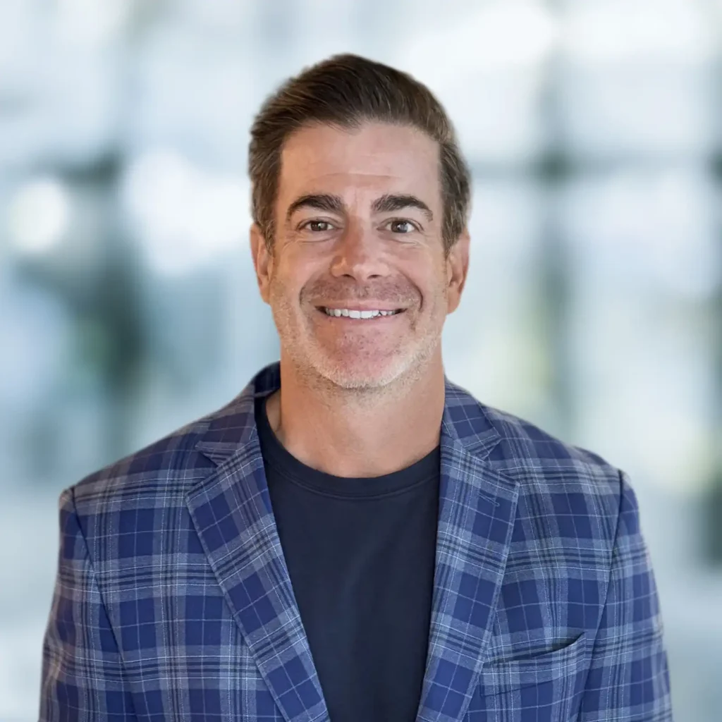 A man with short brown hair, wearing a blue plaid blazer over a dark shirt, smiles at the camera against a blurred indoor background.